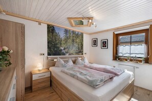 A bedroom with a wooden ceiling, a bed with a floral bedspread, a nightstand with a lamp, a window with a view of trees, and a framed picture on the wall. - Apartment 'Steinbühlschlössle' with Shared Terrace, Shared Garden and Wi-Fi (Schönwald im Schwarzwald)