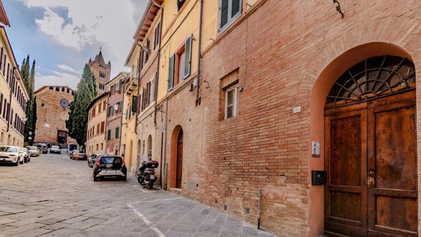 A cobblestone street lined with historic buildings, a prominent brick building with a large wooden door, and a tower in the background. - GuestHost - Porta Romana Charme Apartment (Siena)