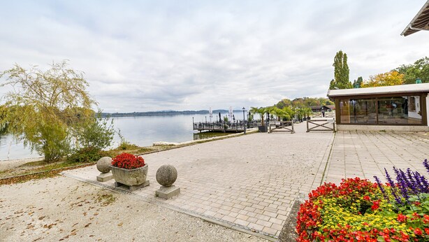 A waterfront promenade with a paved walkway, stone planters with flowers, a dock, and a building with a glass facade.
