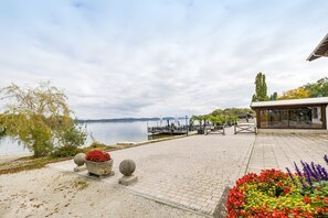 A waterfront promenade with a paved walkway, stone planters with flowers, a dock, and a building with a glass facade.