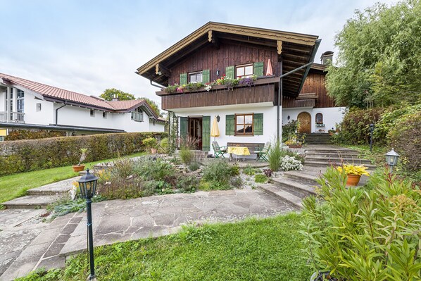 A traditional chalet with a stone pathway, a wooden balcony with plants, and a bench outside.