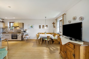 A kitchen with wooden cabinets, a dining table with chairs, a television on a wooden cabinet, and a window with curtains. - Apartment 'Kauderer - Wohnung 1' with Private Terrace, Shared Garden and Wi-Fi (Waging am See)
