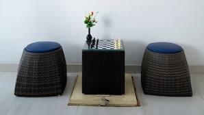 Una habitación con un tablero de ajedrez sobre una mesa de madera, dos taburetes de mimbre y un cuadro floral en la pared.