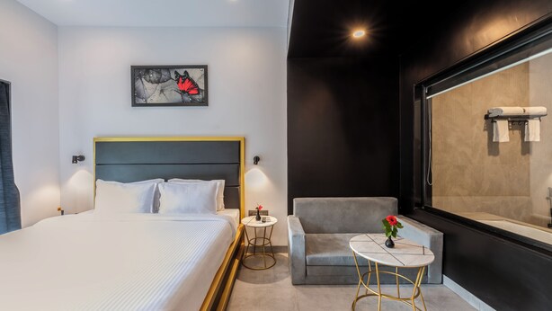 Una habitación de hotel moderna con una cama grande, una mesita redonda pequeña, un sillón gris y un ventanal con vistas a una bañera.