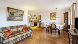 A living room with a sofa, dining table, chairs, a TV, and a kitchen in the background. - Apartment 'Kauderer - Wohnung 2' with Private Terrace, Shared Garden and Wi-Fi (Waging am See)