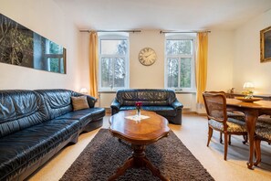 A living room with a black leather sofa, a wooden coffee table, a dining table with chairs, and a wall clock. - Apartment 1 – Nature Retreat at the Resort near Lychen (Lychen)