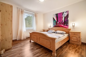 Ein Schlafzimmer mit einem hölzernen Bett, einem Fenster mit Vorhängen, einem Nachttisch und einer Wandlampe.