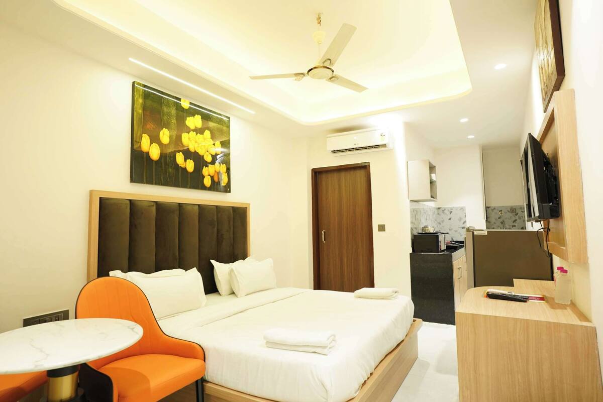 A hotel room with a bed, orange chairs, a small table, a ceiling fan, an air conditioning unit, a kitchen area, and a TV.