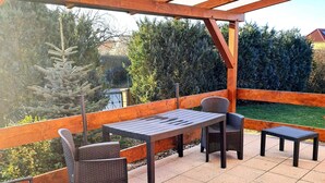 An outdoor patio with wooden beams, a table, chairs, and a view of the surrounding greenery.