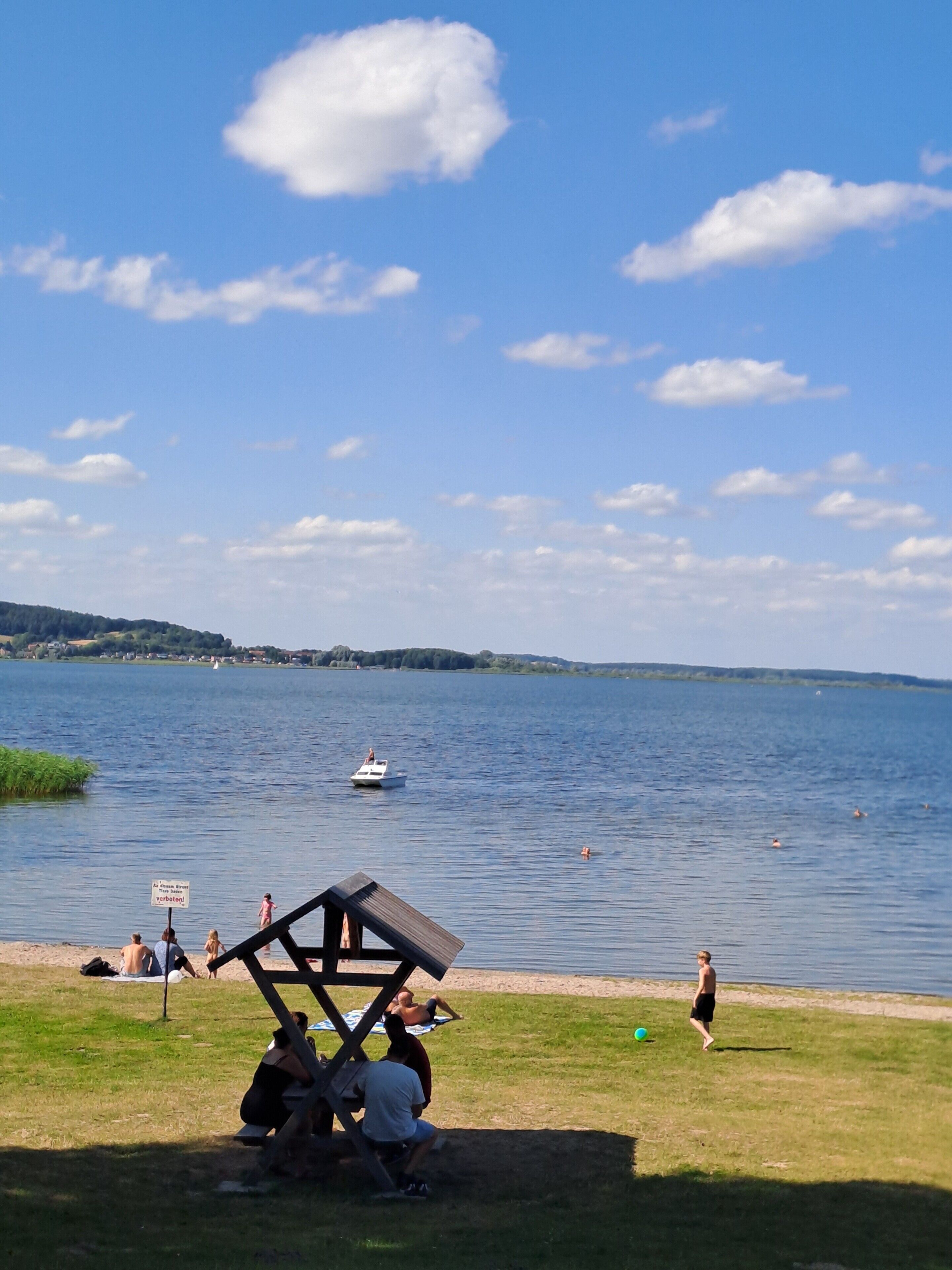 Eine Uferlandschaft mit einem Boot, Menschen entspannen im Gras und einem klaren Himmel.