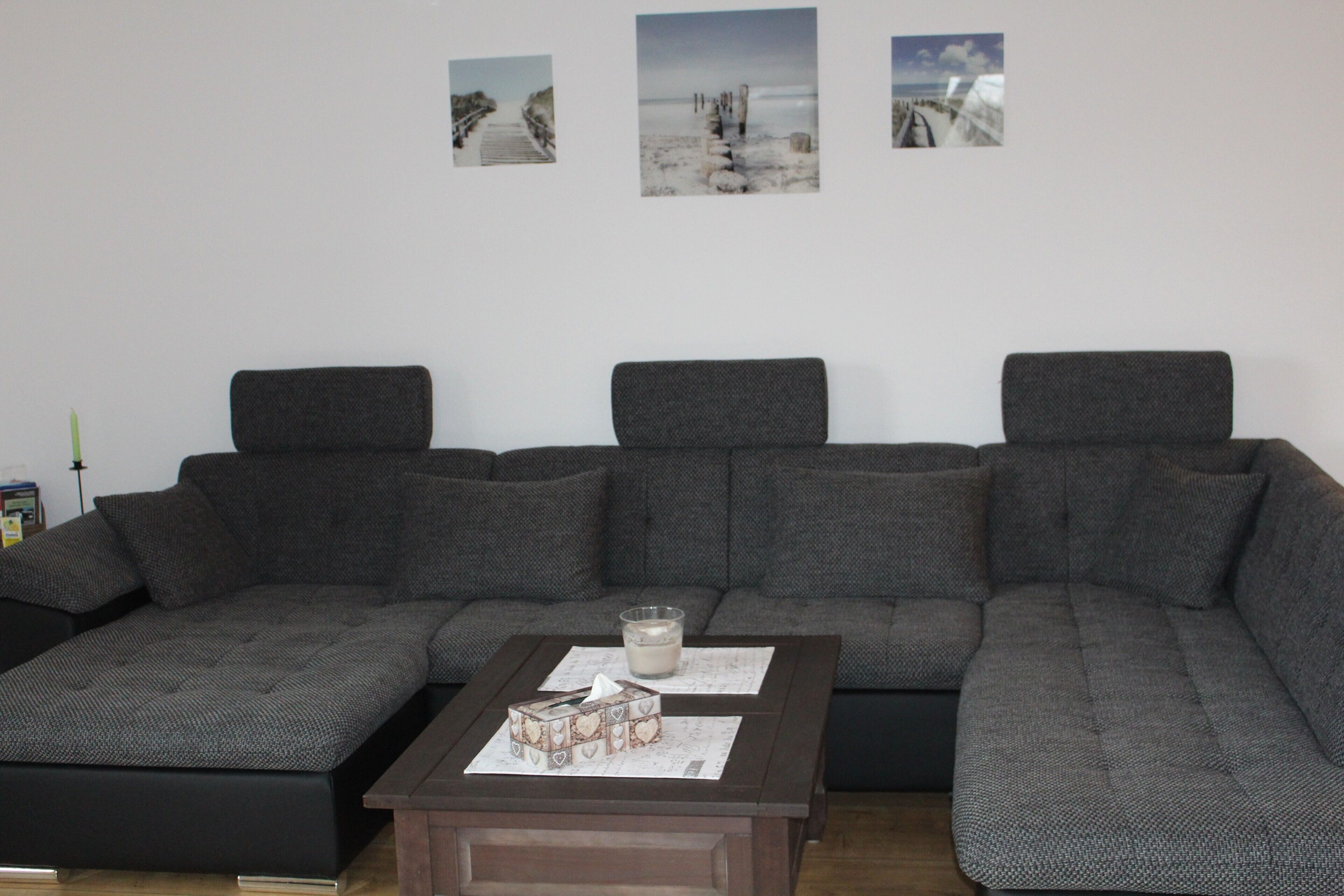 Uma sala de estar moderna com um sofá seccional, uma mesa de centro com uma vela e uma caixa, e três quadros na parede.