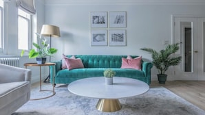 Ein Wohnzimmer mit einem petrolfarbenen Sofa, einem runden Couchtisch und einer Stehlampe. An der Wand hängen gerahmte Bilder und auf dem Boden steht ein Blumentopf.