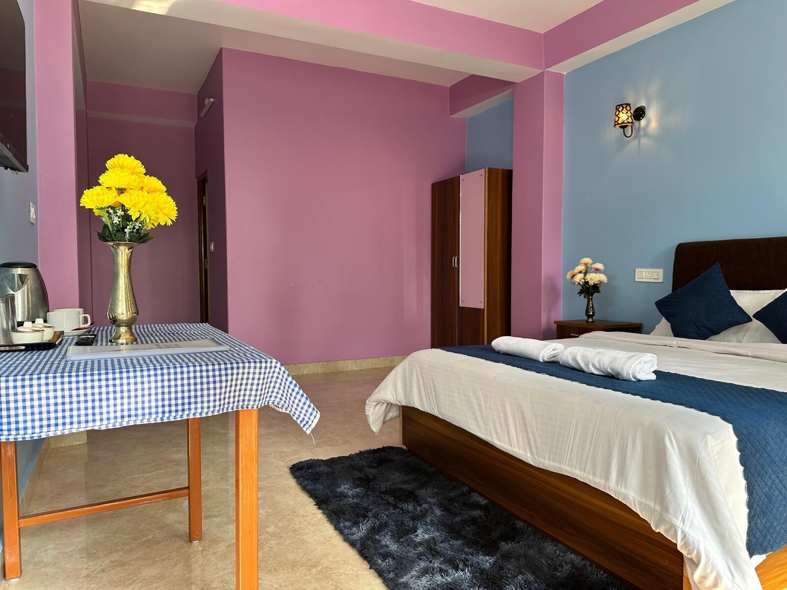 A hotel room with a bed, a table with a checkered tablecloth, a vase with yellow flowers, a nightstand with a lamp, and a dark rug.