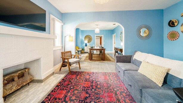 Un salón con pared azul, sofá gris, alfombra roja estampada y un comedor con mesa y sillas.