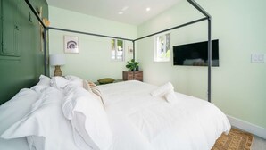 Ein ordentlich bezogenes Bett mit weißen Leinen, eine Nachttischlampe, ein gerahmtes Bild, eine kleine Pflanze und ein Fernseher an der Wand.