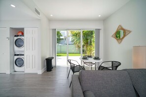 A compact living space with a sofa, a dining table with chairs, a washing machine, and a trash bin.