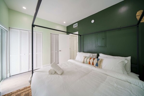 A bedroom with a bed, white linens, a green accent wall, and a window with blinds.