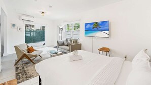 A modern hotel room with a large bed, a seating area with a sofa and armchair, a flat-screen TV, and a view of the outdoors through the glass door. - Designer Studio | Close to Wynwood | Design District  | Downtown (Miami)