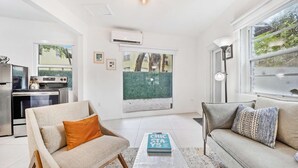 A modern living room with a sofa, armchair, and coffee table. There is a kitchen area with appliances and a large window. - Designer Studio | Close to Wynwood | Design District  | Downtown (Miami)