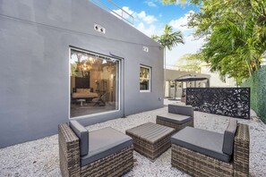 Un espacio moderno para sentarse al aire libre con mobiliario de mimbre y un muro de cristal con vistas a un dormitorio.
