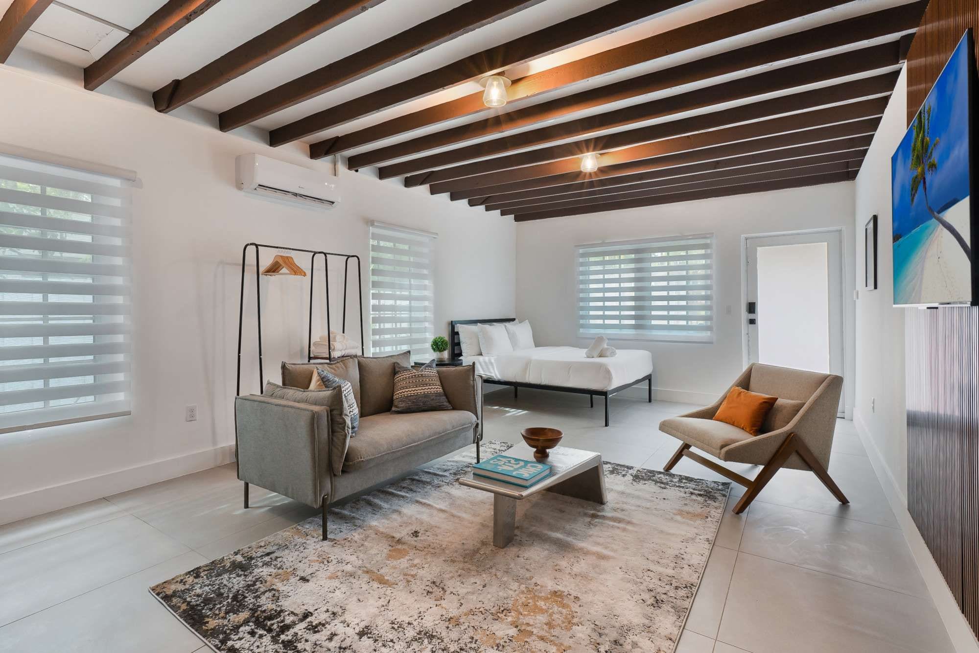 Un salon moderne avec un canapé, un fauteuil et une table basse, un lit avec une literie blanche et une grande fenêtre équipée de stores.