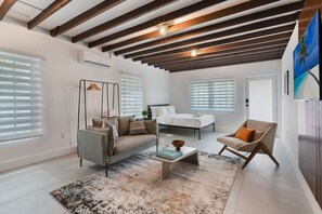 A modern living room with a sofa, armchair, and coffee table, a bed with white bedding, and a large window with blinds. - Designer Studio | Close to Wynwood | Design District  | Downtown (Miami)