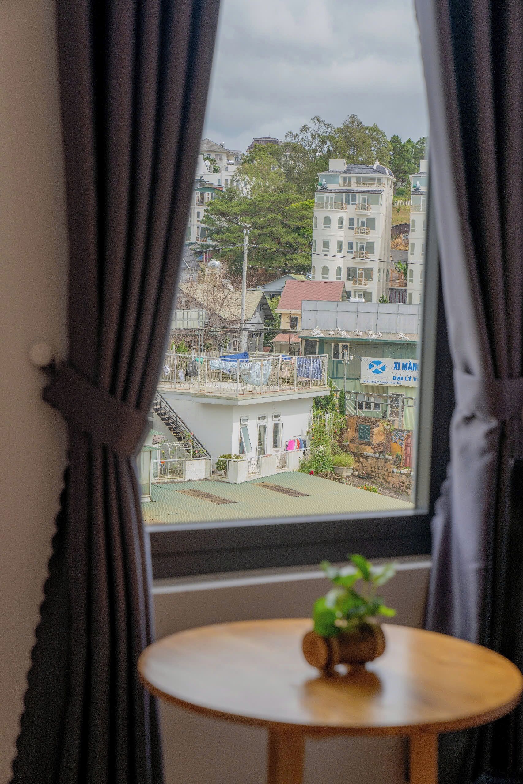 Habitación con ventana que da a una zona residencial, una mesita redonda con una planta y cortinas corridas a un lado.