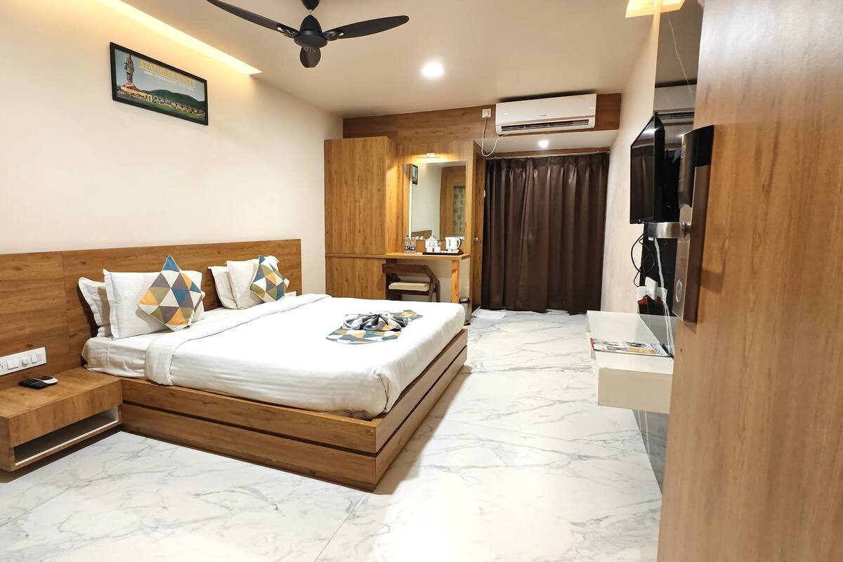 A modern bedroom with a wooden headboard, a bed with patterned pillows, a bedside table with a remote control, a wooden desk with a chair, a mirror, and an air conditioning unit.