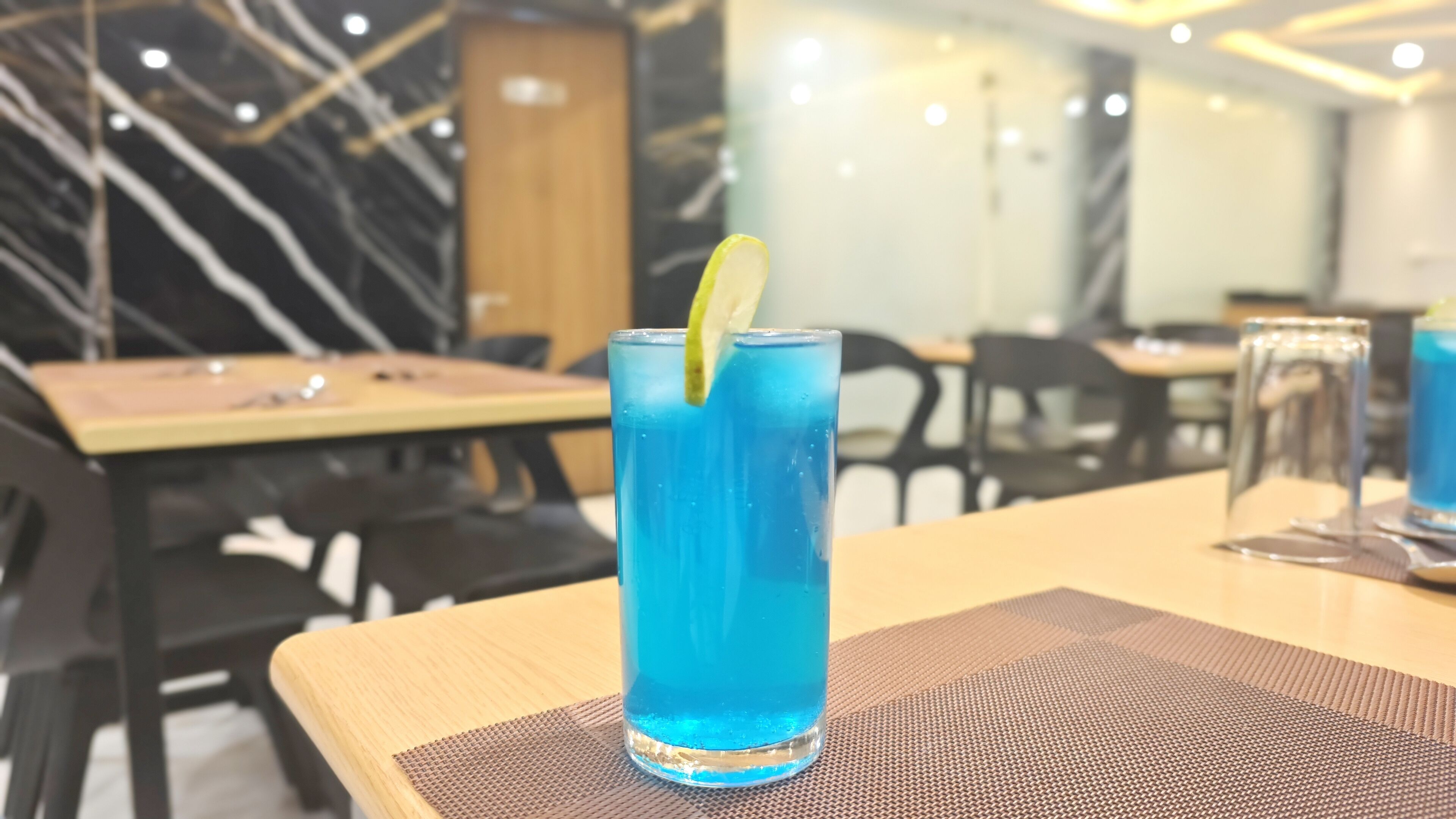 L’intérieur d’un restaurant, avec une table sur laquelle se trouve une boisson bleue décorée d’une tranche de citron vert, un mur en marbre noir et blanc, et une porte en bois en arrière-plan.