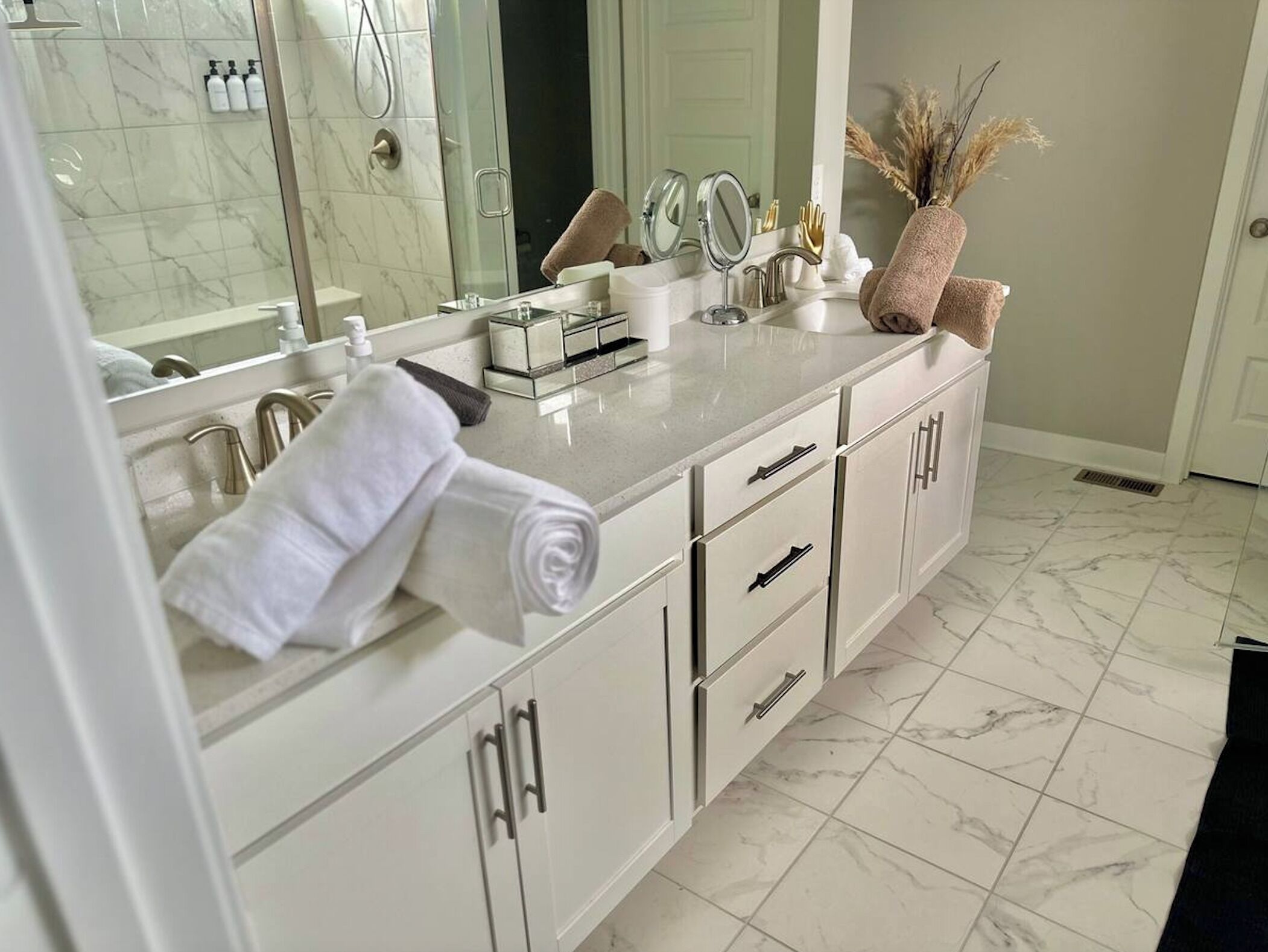 A modern bathroom with a large mirror, white cabinets, and a marble floor.