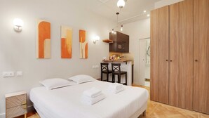 A modern bedroom with a large bed, a wooden wardrobe, a small kitchenette, and abstract wall art. - Cozy studio - 2P - Gare de l'Est (Paris)