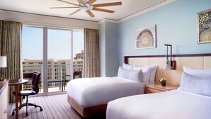 A hotel room with two beds, a desk, a chair, a ceiling fan, and a large window with curtains.