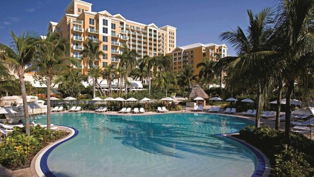 A resort pool with lounge chairs and umbrellas, surrounded by palm trees and a hotel building in the background.