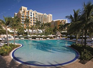 Piscina de un resort con sillones de descanso y sombrillas, rodeada de palmeras y un edificio de hotel al fondo.