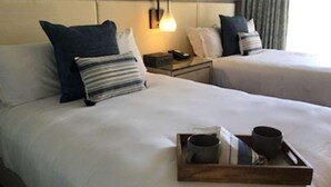 A hotel room with two beds, each with white bedding and blue pillows. There is a bedside table with a lamp and a phone. A tray with a cup and a mug is on the bed.