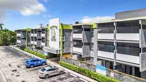 Un edificio moderno con fachada verde y blanca, varios balcones y un estacionamiento con dos coches.