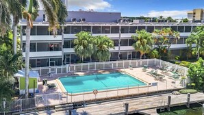 Una piscina en la azotea con un deck de madera, rodeada de palmeras y una valla blanca.