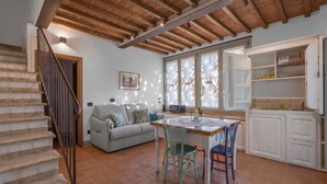 A spacious living area with a wooden staircase, a dining table with chairs, a sofa, and a kitchen area with cabinets and a window with curtains. - La Capanna, Borgo Campassini, Monteriggioni (Monteriggioni)