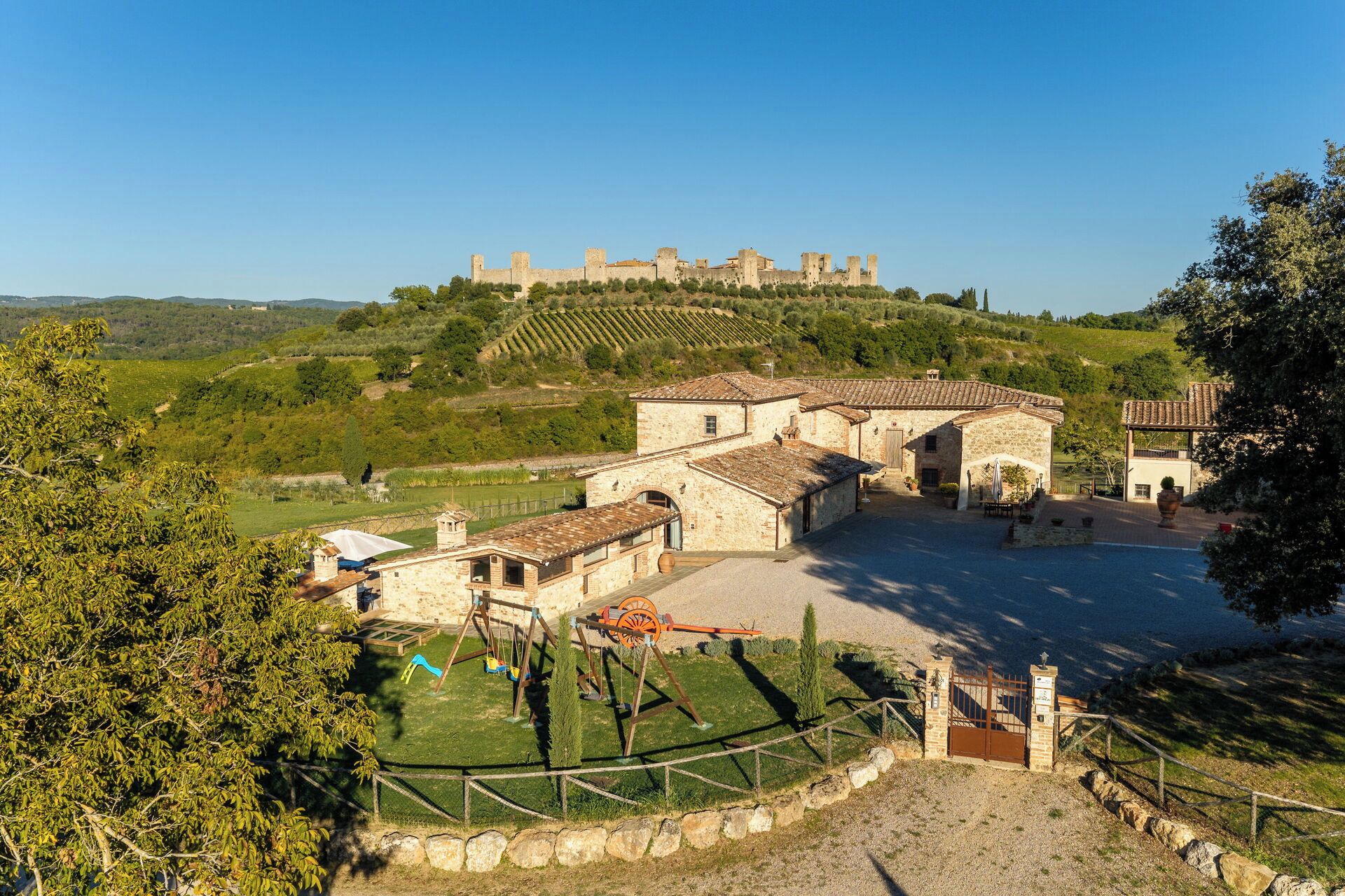 A stone building with a courtyard, surrounded by trees and a view of a hill with ruins.