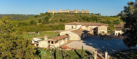 A stone building with a courtyard, surrounded by trees and a view of a hill with ruins.