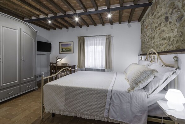 A bedroom with a bed, a desk with a lamp, a wardrobe, and a stone wall.