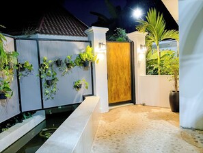 Une entrée fermée par un portail et une porte en bois, agrémentée de plantes en pot et d’un chemin en béton menant à une zone piscine.