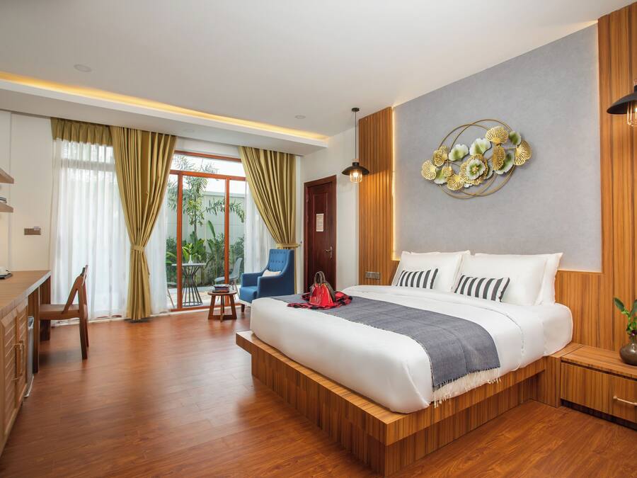 A modern hotel room with a large bed, wooden flooring, a desk with a chair, a blue armchair, and a view of greenery through the window.