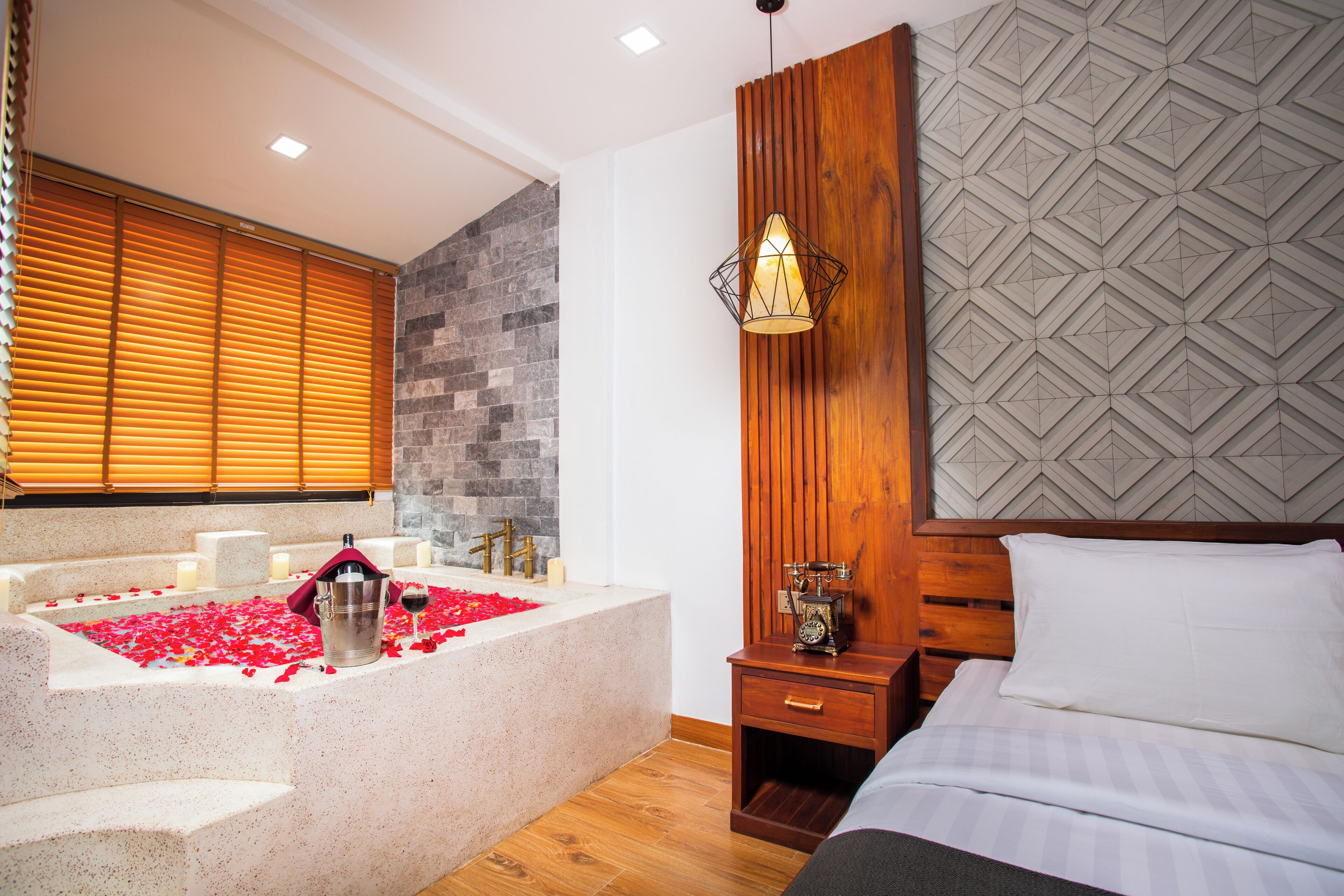 A modern bedroom with a large bed, a wooden nightstand, a stone bathtub with rose petals, and a geometric-patterned wall.