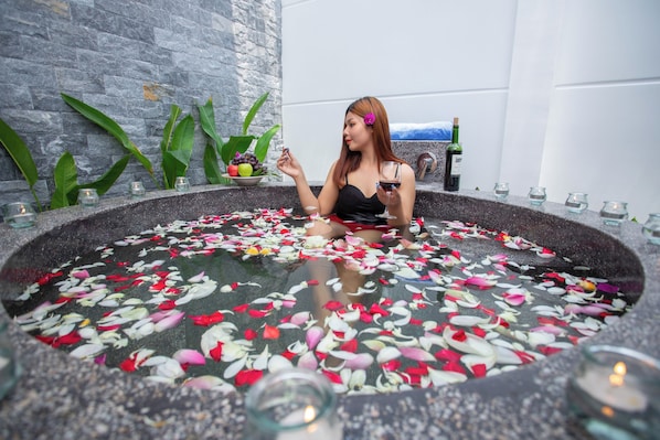 A woman sitting in a stone bathtub filled with water and rose petals, surrounded by candles and greenery.