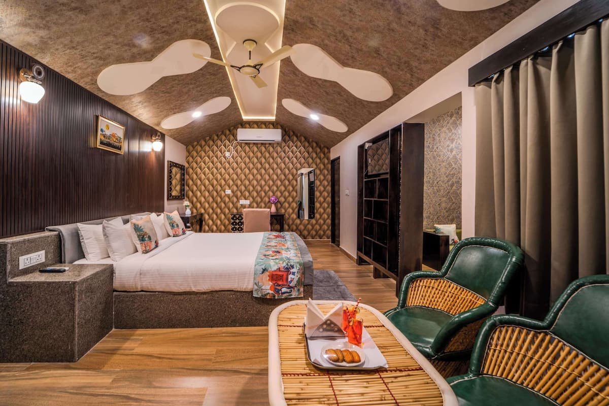 A hotel room with a large bed, a sofa, two green chairs, a small table with a tray of snacks, and a ceiling fan.
