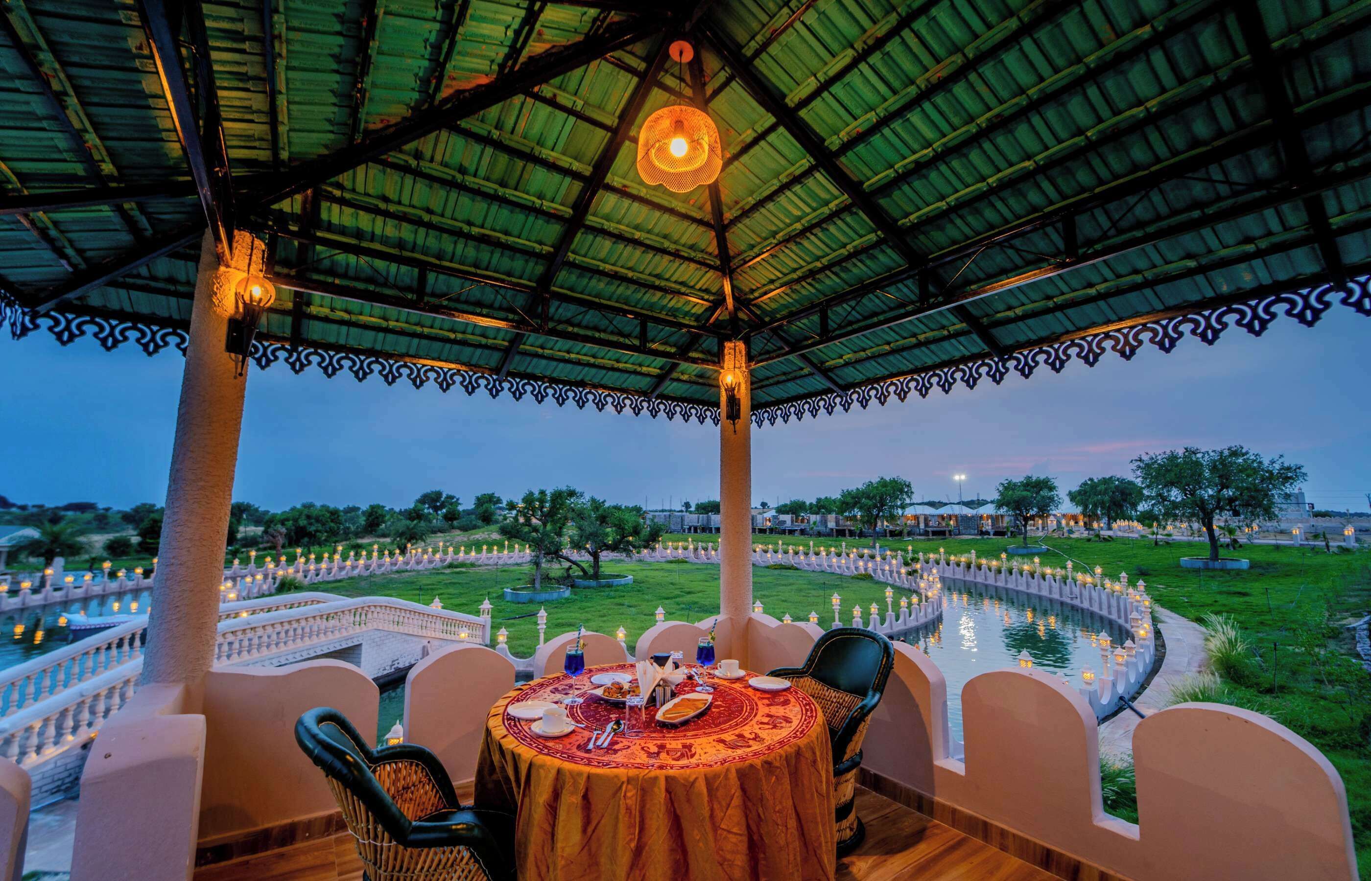 An outdoor dining area with a table set for two, surrounded by chairs, under a covered structure with a patterned roof.
