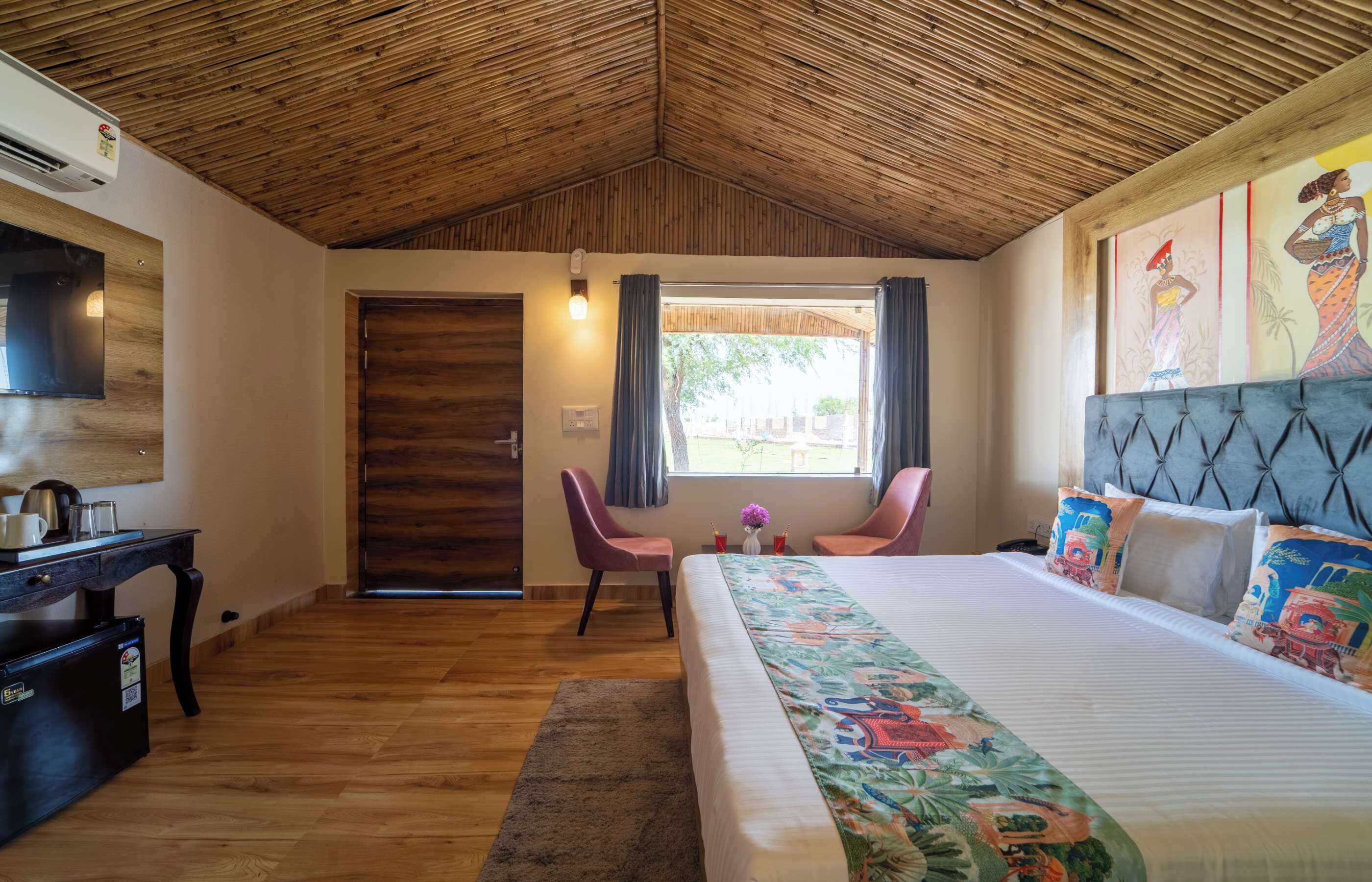 A bedroom with a wooden ceiling, a bed with colorful bedding, a desk with a kettle, and a TV on the wall.