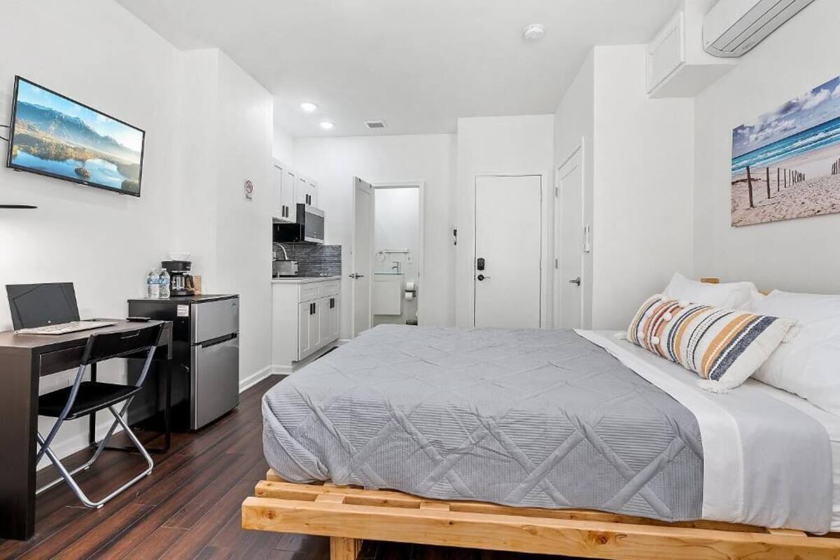 A bedroom with a bed, a desk, a chair, a kitchenette, and a TV on the wall.