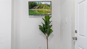 A room with a television mounted on the wall displaying a scenic landscape, a potted plant, and a white door. - Ututu Stay West (Philadelphia)